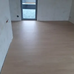 Pokój w trakcie wykańczania, z nową podłogą laminowaną o wzorze drewna, ściany przygotowane do malowania, widoczne gniazdka elektryczne i szpachlowane ubytki, okno z widokiem na sąsiedni budynek.