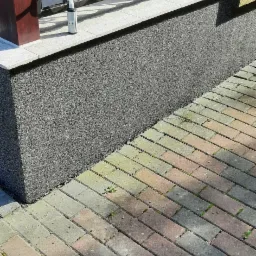 Szara elewacja z fakturą baranka przy balkonie z metalową balustradą, obok kostka brukowa i butelka impregnatu.