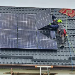 Montaż paneli słonecznych na dachu pokrytym blachodachówką; pracownik w kasku i uprzęży zabezpieczającej układa kolejny panel, w tle dywanik zabezpieczający.