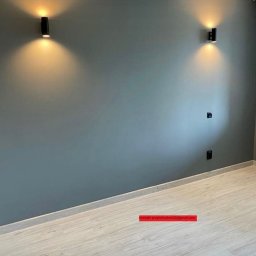 Przytulny Dom - Szara ściana z dwoma czarnymi lampami ściennymi LED, oświetlającymi przestrzeń ciepłym światłem, oraz widocznymi gniazdkami elektrycznymi i fragmentem podłogi z paneli.