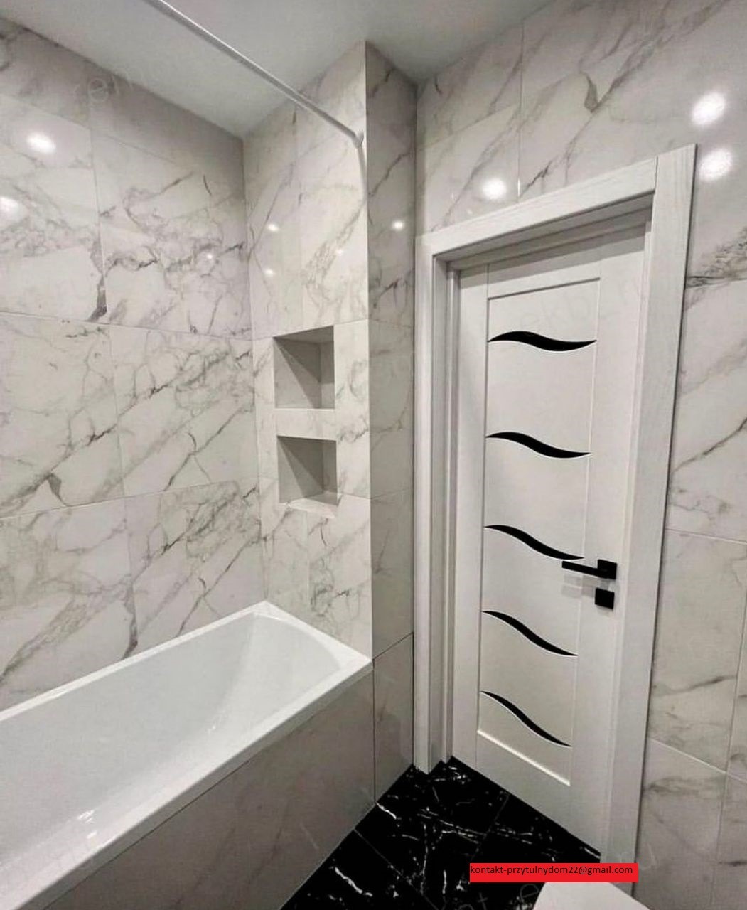 Wykończona łazienka z wanną, białą armaturą i marmurowymi płytkami ściennymi z wnękami, czarna podłoga, białe drzwi z czarnymi, falistymi zdobieniami i klamką.