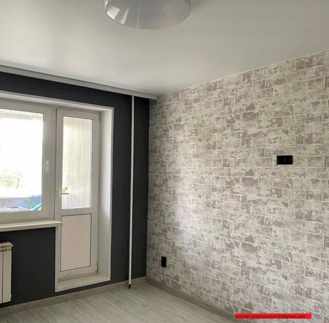 Pokój po remoncie z tapetą imitującą cegłę, ciemnoszara wnęka okienna, białe drzwi balkonowe i grzejnik. Widoczna lampa sufitowa i kontakt.
