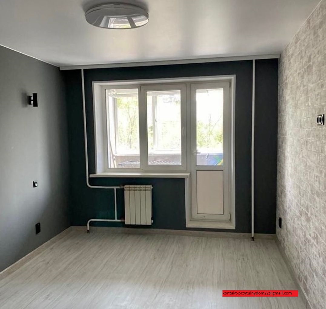 Pomalowane na ciemny grafit pokój z jasną podłogą imitującą drewno, białe okno balkonowe z grzejnikiem pod spodem, jedna ściana pokryta tapetą imitującą cegłę.