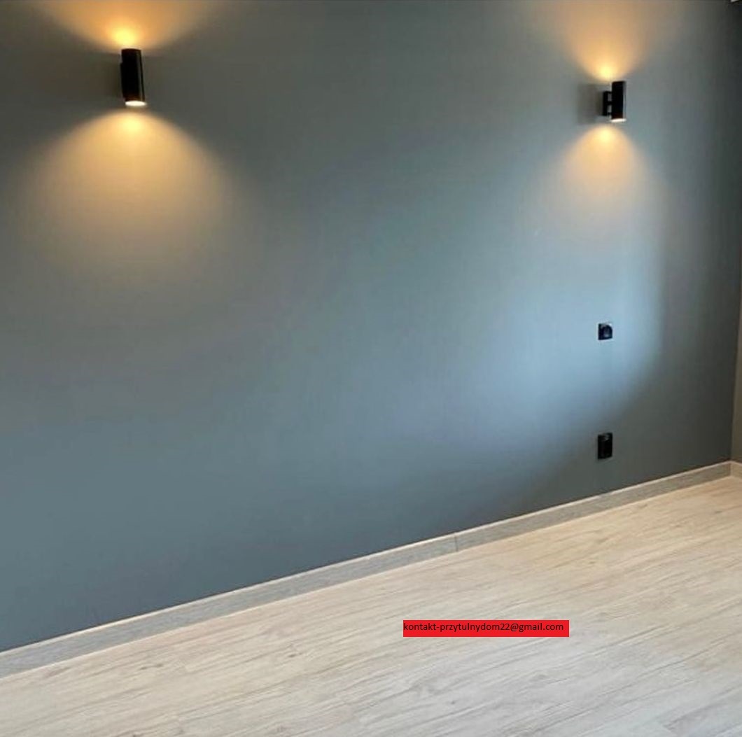 Szara ściana z dwoma czarnymi lampami ściennymi LED, oświetlającymi przestrzeń ciepłym światłem, oraz widocznymi gniazdkami elektrycznymi i fragmentem podłogi z paneli.