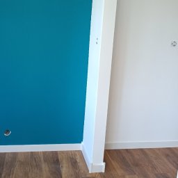 RAW-WYKONCZENIE WNETRZ Adam Rawski - Pomalowany na turkusowo fragment ściany w rogu pomieszczenia, widoczna podłoga z paneli imitujących drewno i biała kolumna z listwą przypodłogową.