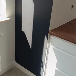 Ciemnografitowa ściana w pokoju, oświetlona padającym światłem słonecznym, widoczny fragment białej szafki z drewnianym blatem i jasne płytki podłogowe.