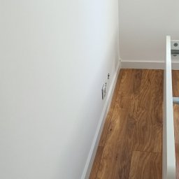 RAW-WYKONCZENIE WNETRZ Adam Rawski - Świeżo pomalowana, biała ściana w rogu pokoju, widoczne gniazdka elektryczne i listwa przypodłogowa, podłoga wyłożona panelami imitującymi drewno.