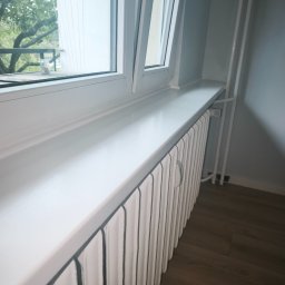 RAW-WYKONCZENIE WNETRZ Adam Rawski - Biały, odnowiony grzejnik pod oknem z widokiem na zieleń. Świeżo pomalowany, z nowym parapetem, na tle drewnianej podłogi i jasnych ścian.