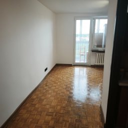 RAW-WYKONCZENIE WNETRZ Adam Rawski - Jasny pokój z białymi ścianami i lśniącym, kwadratowym parkietem. W tle drzwi balkonowe z widocznym drewnianym krzesłem i białym grzejnikiem pod oknem.