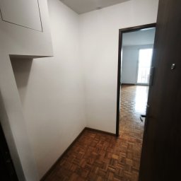 RAW-WYKONCZENIE WNETRZ Adam Rawski - Czysty korytarz z białymi ścianami, parkietem w kwadratowy wzór i nowoczesnymi reflektorami. Widoczne częściowo otwarte ciemne drzwi i biała zabudowa z panelem.