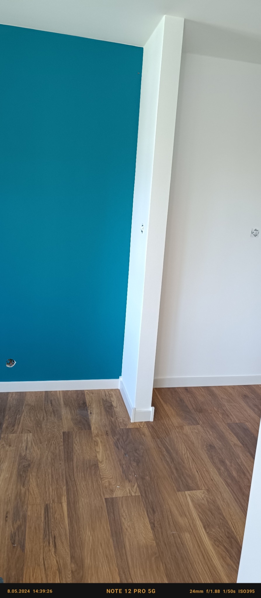 Pomalowany na turkusowo fragment ściany w rogu pomieszczenia, widoczna podłoga z paneli imitujących drewno i biała kolumna z listwą przypodłogową.