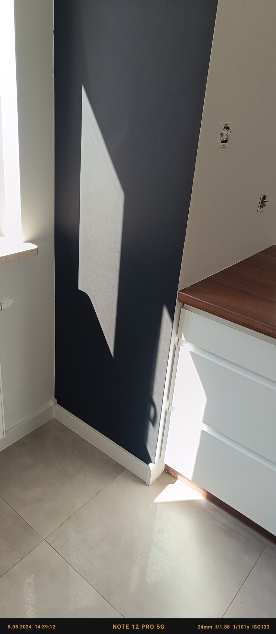 Ciemnografitowa ściana w pokoju, oświetlona padającym światłem słonecznym, widoczny fragment białej szafki z drewnianym blatem i jasne płytki podłogowe.