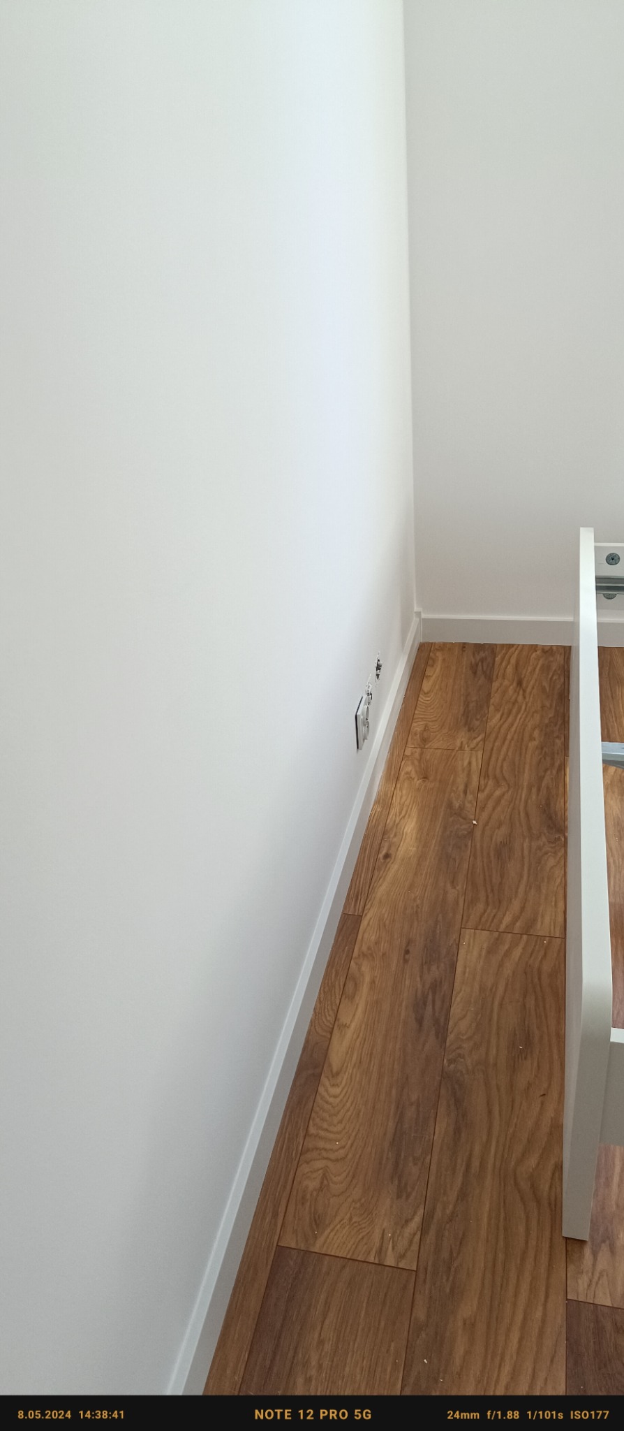 Świeżo pomalowana, biała ściana w rogu pokoju, widoczne gniazdka elektryczne i listwa przypodłogowa, podłoga wyłożona panelami imitującymi drewno.