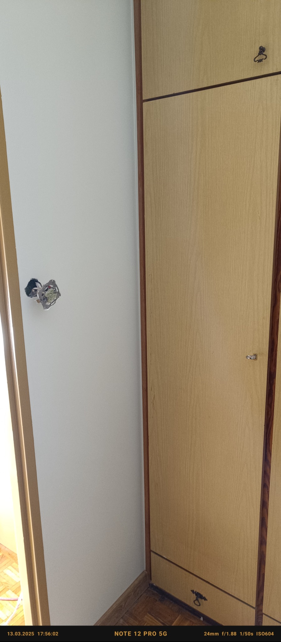 Biała ściana z wystającą puszką elektryczną przed montażem gniazdka, obok drewniana szafa.