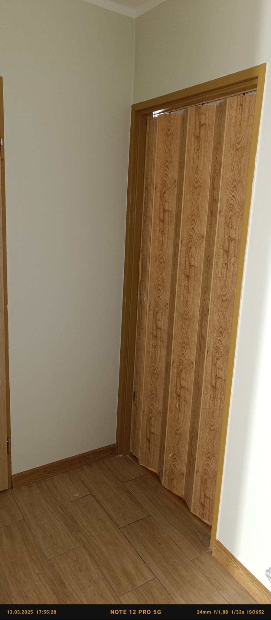 Jasne wnętrze z panelami ściennymi imitującymi drewno i składanymi drzwiami w odcieniach brązu, widoczne fragmenty podłogi z paneli.