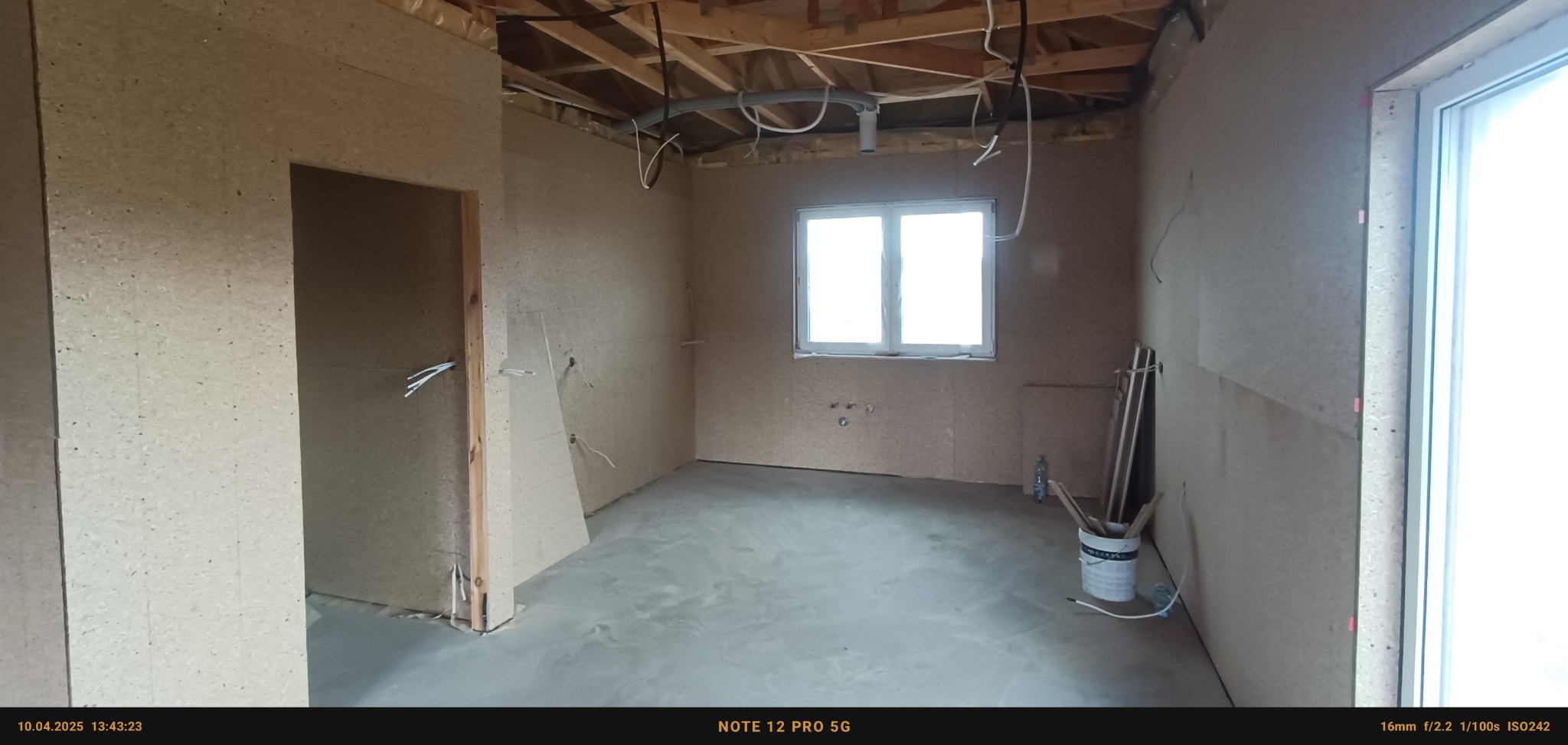 Wnętrze w trakcie prac przygotowawczych: ściany obłożone płytami OSB, widoczne instalacje elektryczne i hydrauliczne, surowy stan podłogi, okno w centralnej części, wiadro i narzędzia w rogu...