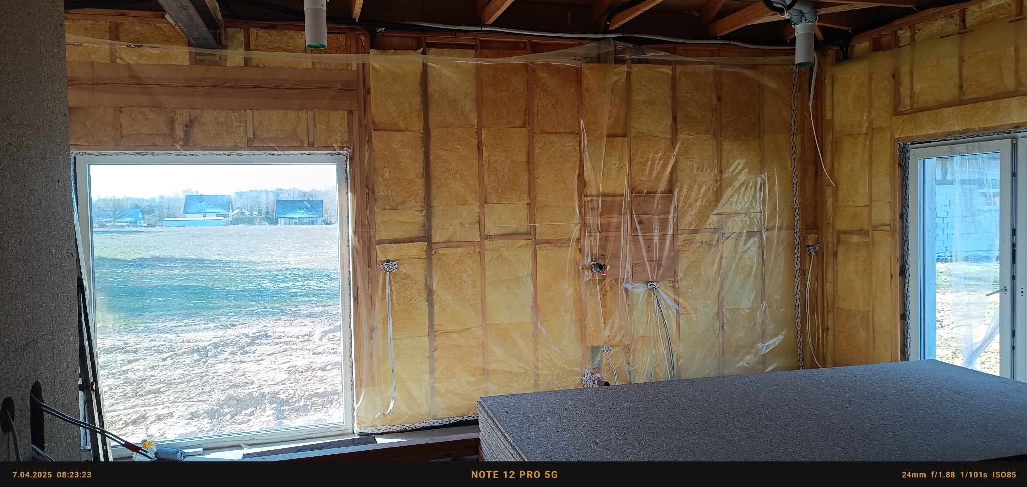 Wnętrze domu szkieletowego w trakcie izolacji, widoczna paroizolacja na ścianach z wełną mineralną, okno z widokiem na krajobraz, przygotowane płyty OSB na pierwszym planie.