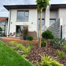 Nowoczesny ogród przydomowy z drewnianym tarasem, meblami ogrodowymi, ozdobnymi trawami w donicach, młodymi drzewami podpartymi palikami i rabatami wysypanymi korą.