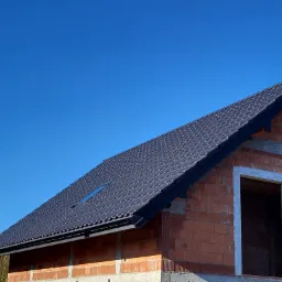 Nowy, grafitowy dach z dachówki ceramicznej na budynku w trakcie budowy, widoczne okno dachowe i orynnowanie, błękitne niebo w tle.