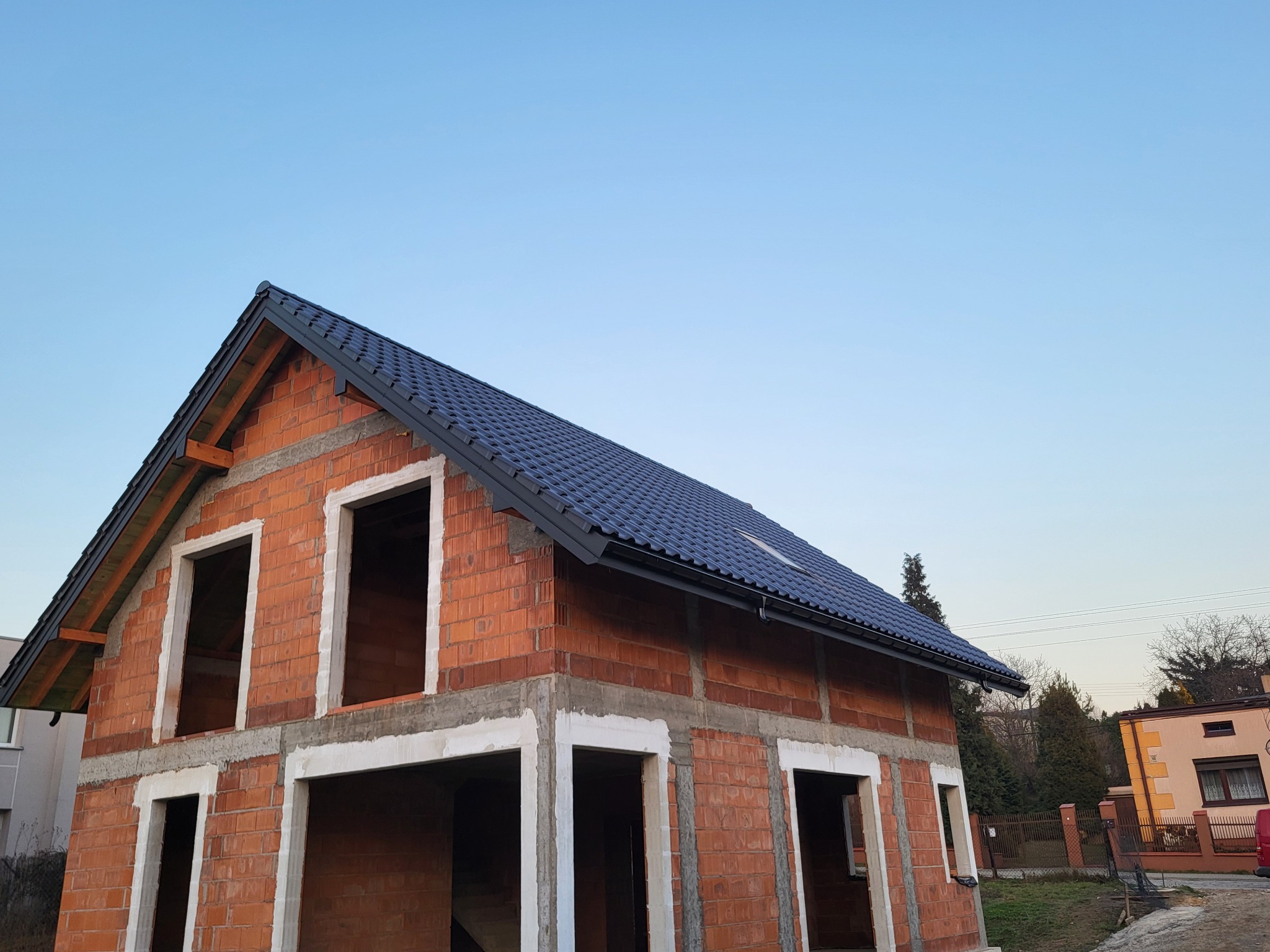 Surowy budynek w trakcie budowy z częściowo ukończonym dachem z ciemnej dachówki, widoczne okna bez ram, ceglana konstrukcja i błękitne niebo w tle.