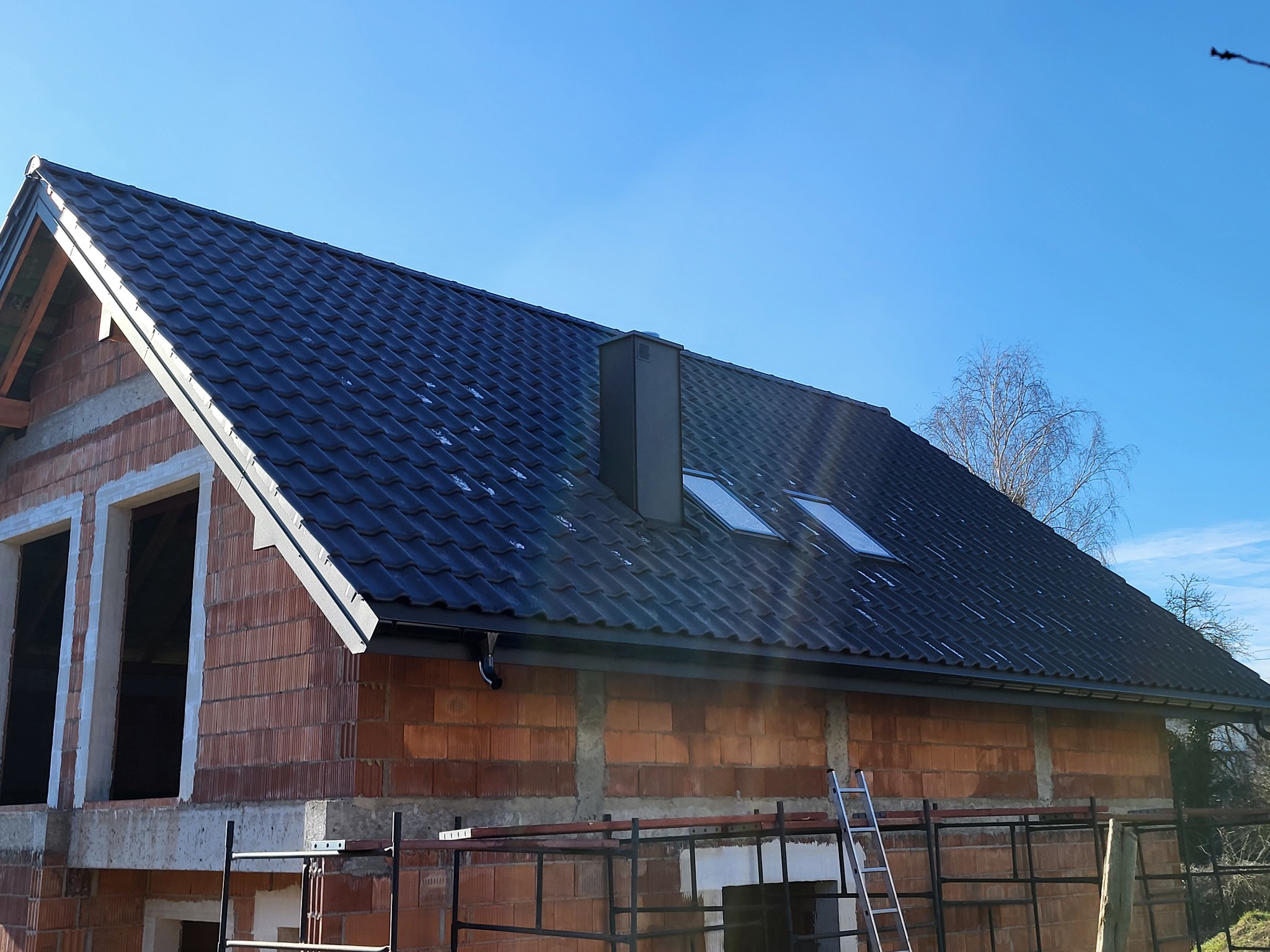 Nowy, ciemny dach z oknami dachowymi i kominem na surowym budynku z cegły, widok z dołu na tle błękitnego nieba.