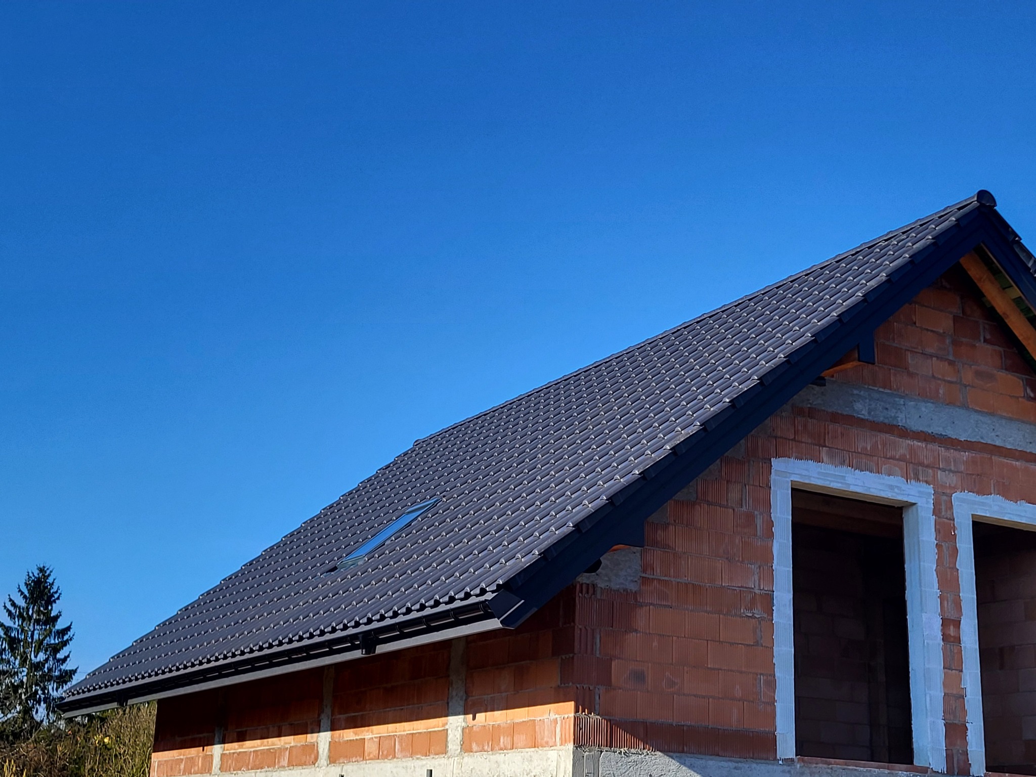 Nowy, grafitowy dach z dachówki ceramicznej na budynku w trakcie budowy, widoczne okno dachowe i orynnowanie, błękitne niebo w tle.