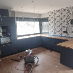 Wnętrze kuchni w trakcie remontu: ciemne meble kuchenne z drewnianym blatem, ściana z tapetą w liście, widoczne narzędzia, drabina i laser poziomujący.