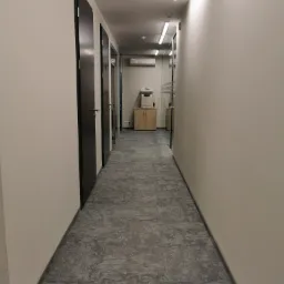 Długi korytarz biurowy z szarą wykładziną dywanową, białymi ścianami i ciemnymi drzwiami, zakończony szafką z drukarką i klimatyzacją.
