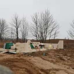 Wznoszenie ścian parteru domu z bloczków betonu komórkowego na fundamencie, widoczne narzędzia budowlane i materiały, krajobraz z drzewami bez liści w tle.