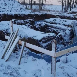 Zasypana śniegiem wykopana ziemia pod fundamenty z drewnianym szalunkiem, widok na zaśnieżone pole i drzewa bez liści w tle.