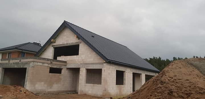 Surowy dom murowany z ciemnym dachem w trakcie budowy, widoczne nieotynkowane ściany z pustaków, sterta piasku z przodu, pochmurne niebo w tle.