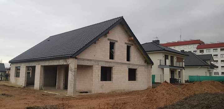 Surowy, niewykończony dom jednorodzinny z czarną dachówką, widoczne otwory okienne i drzwiowe, fundamenty częściowo zasypane ziemią, w tle inne budynki.