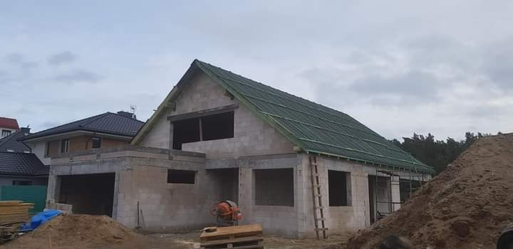 Surowy stan domu jednorodzinnego z pustaków, z zielonym dachem, widoczne rusztowanie i sprzęt budowlany na palecie, w tle inne budynki.