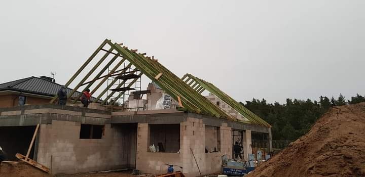 Budowa domu w trakcie wznoszenia więźby dachowej, widoczne ściany z bloczków, rusztowania i pracownicy na dachu, w tle las i sąsiedni dom.