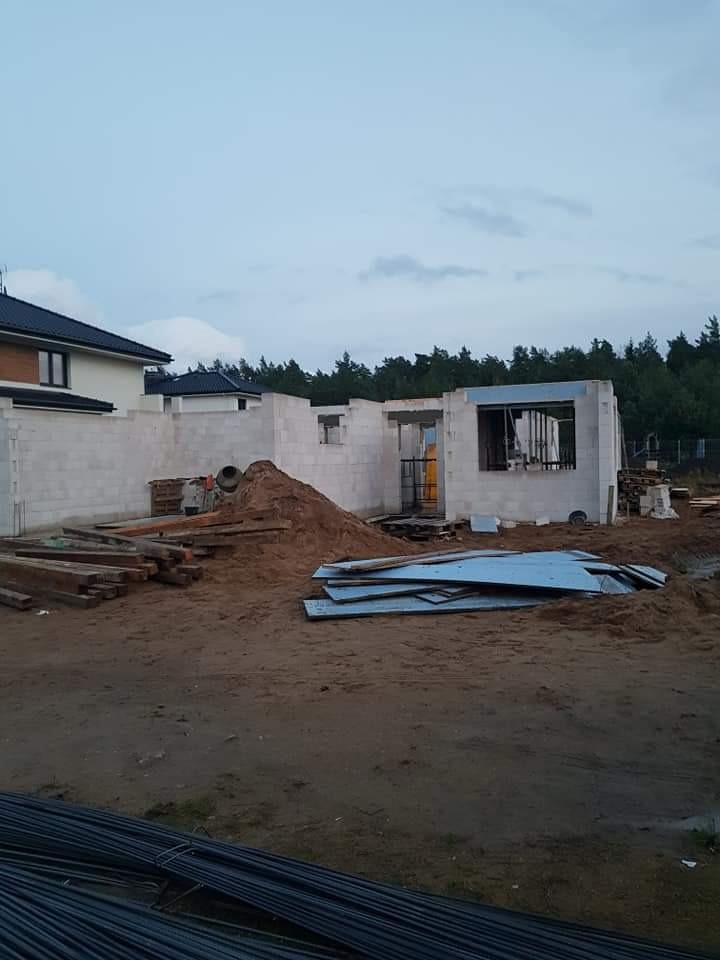 Wznoszenie ścian domu z bloczków betonowych, widoczne sterty materiałów budowlanych i zbrojenia na placu budowy w pochmurny dzień.