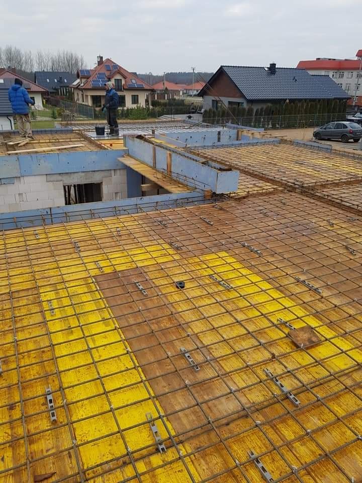 Budowa domu murowanego: zbrojenie stropu na żółtym szalunku, widoczni pracownicy i okoliczne domy w tle.