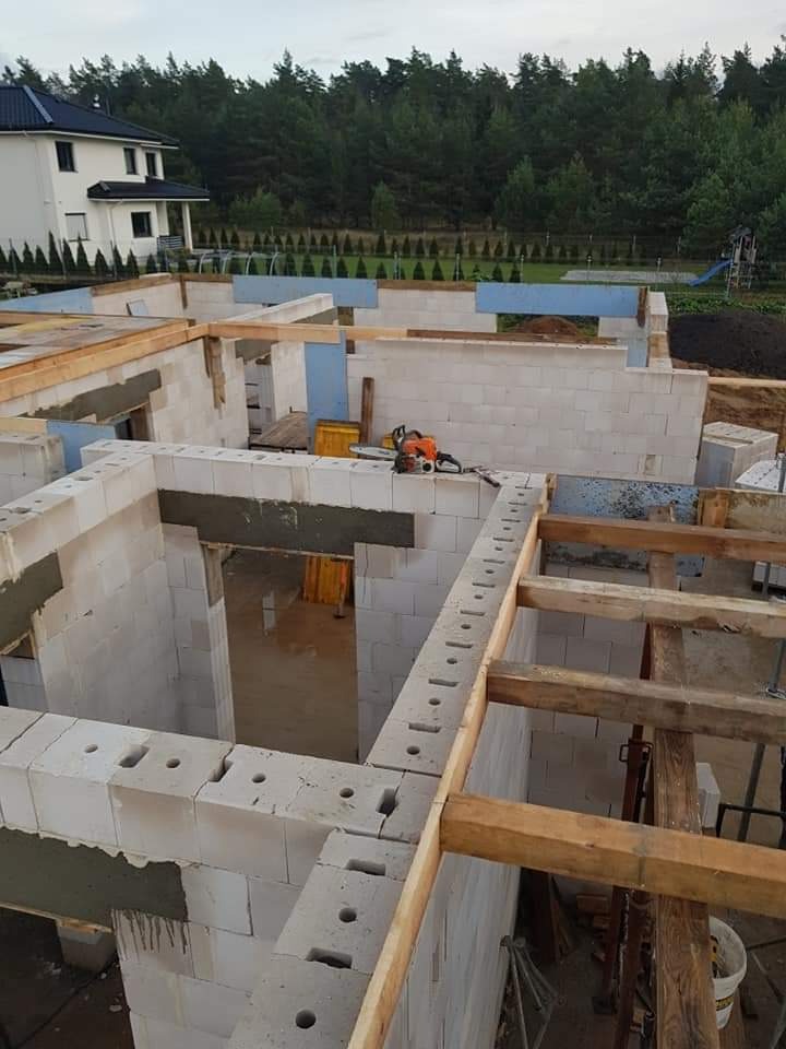 Budowa domu: widok na wznoszone ściany z betonu komórkowego, drewniane wzmocnienia, piła spalinowa na murze, dom w tle.