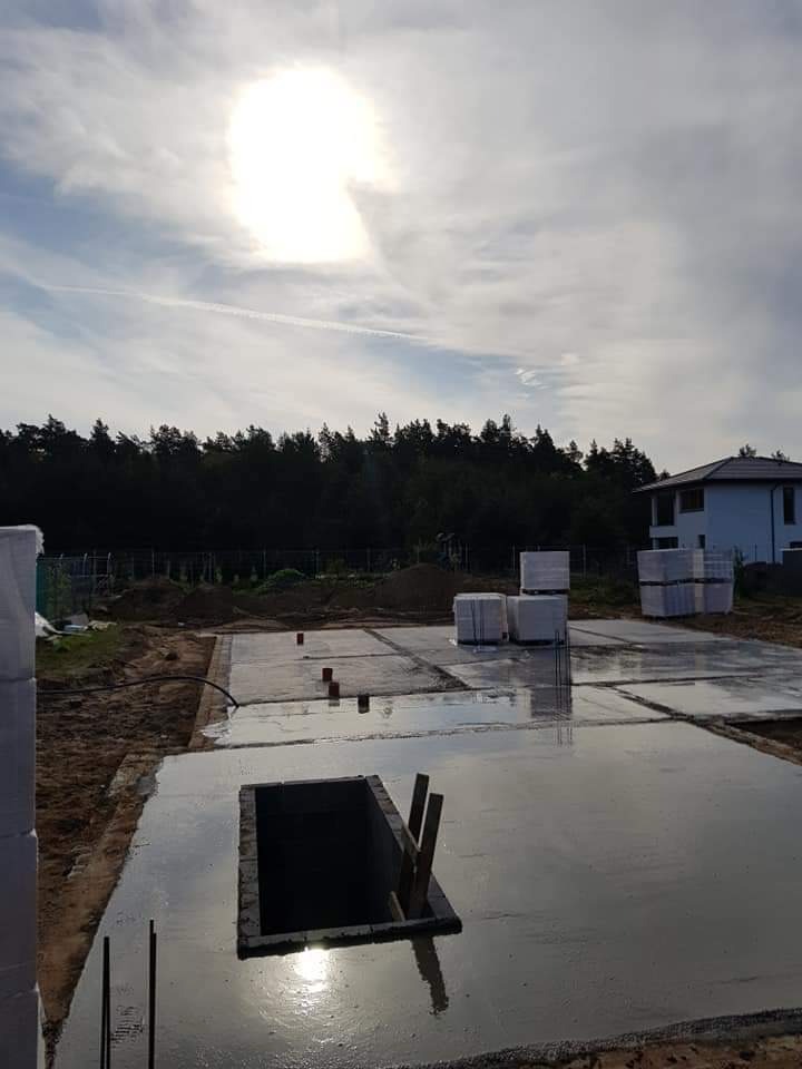 Świeżo wylane, mokre fundamenty pod budynek z widocznym otworem inspekcyjnym, stosami bloczków Ytong i zalesionym terenem w tle.