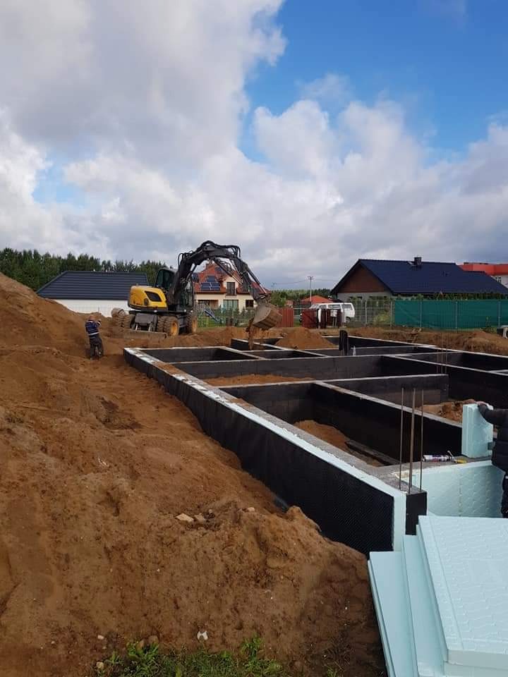Wykop pod fundamenty domu jednorodzinnego, widoczne zbrojenie i izolacja pionowa, w tle koparka i zabudowa mieszkalna.