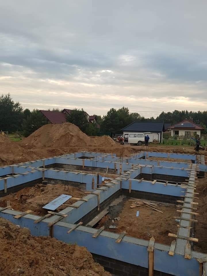 Przygotowane szalunki i izolacja fundamentów pod budowę domu jednorodzinnego, widoczne zbrojenia i elementy konstrukcyjne, w tle hałda ziemi i zabudowa mieszkalna.