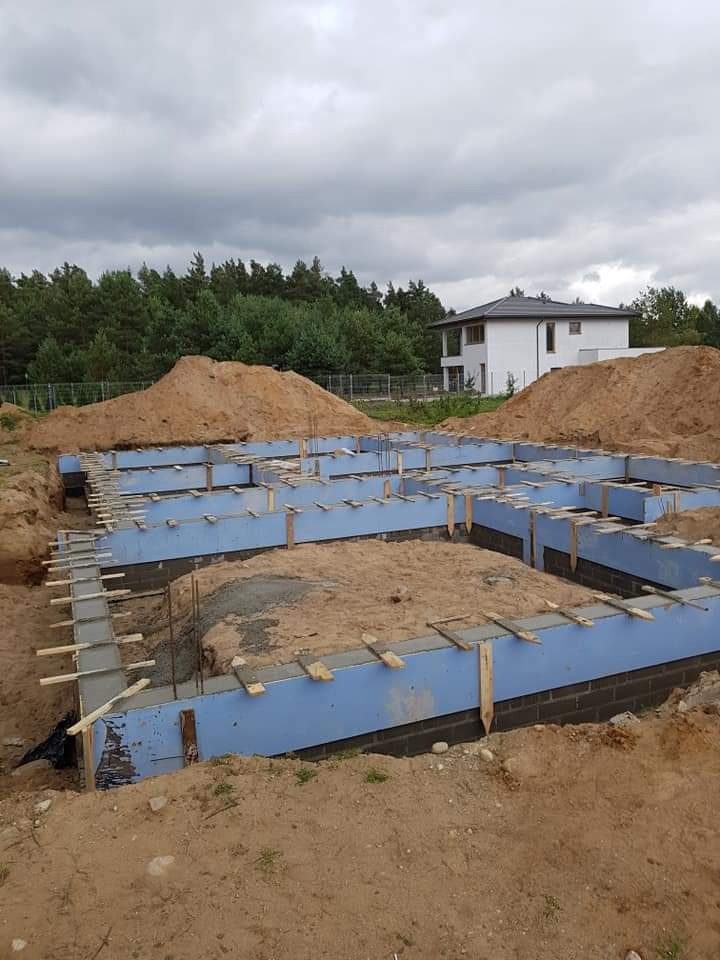 Zalane betonem szalunki fundamentowe z niebieską izolacją, widoczna zbrojenie, piasek zasypowy, w tle dom w budowie i las.