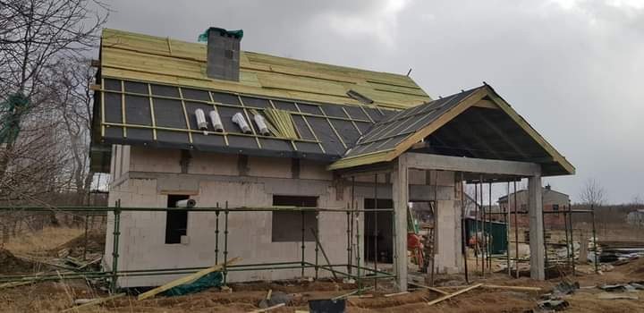 Niedokończony dom z pustaków z widoczną konstrukcją dachu, czarną membraną i częściowo ułożonymi deskami, rusztowania wokół budynku.