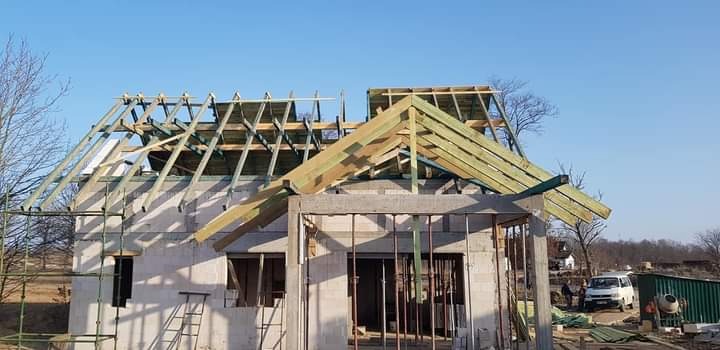 Wznoszenie konstrukcji dachu z drewnianych belek na surowym budynku z białych bloczków, widoczne rusztowania i otoczenie placu budowy.