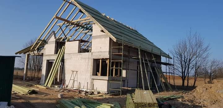 Surowy stan domu z betonu komórkowego z drewnianą konstrukcją dachu, częściowo pokrytą papą, rusztowania przy ścianie bocznej, ułożone deski na ziemi, w tle drzewa bez liści.