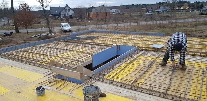 Zbrojenie płyty fundamentowej z otworem na instalacje, pracownik w kraciastej koszuli wiąże drut, w tle zabudowa jednorodzinna.