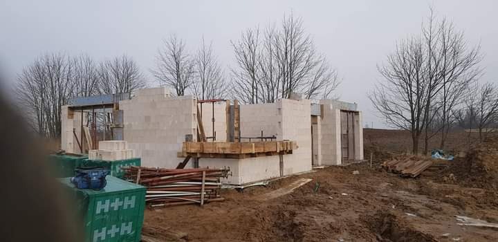 Wznoszenie ścian parterowego domu z bloczków H+H, widoczne drewniane szalunki, teren budowy w pochmurny dzień.