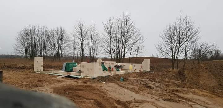 Wznoszenie ścian parteru domu z bloczków betonu komórkowego na fundamencie, widoczne narzędzia budowlane i materiały, krajobraz z drzewami bez liści w tle.