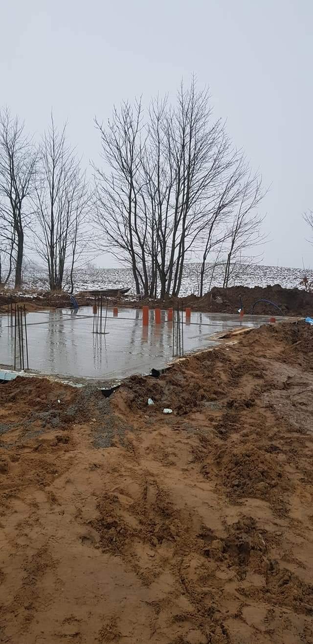 Świeżo wylany fundament pod dom w pochmurny dzień, widoczne wystające pręty zbrojeniowe, pomarańczowe rury i błotniste otoczenie.