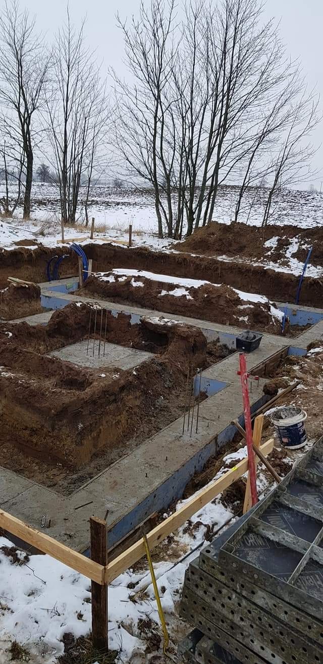 Wykopane fundamenty częściowo zalane betonem, widoczne zbrojenia, szalunki, zasypana śniegiem ziemia i drzewa w tle.