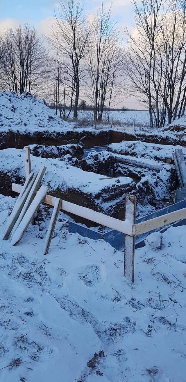 Zasypana śniegiem wykopana ziemia pod fundamenty z drewnianym szalunkiem, widok na zaśnieżone pole i drzewa bez liści w tle.